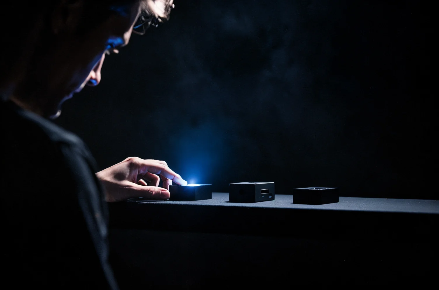 A person in profile examining three small objects laid out on a black bench, one lit hard from the side.